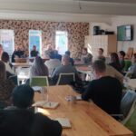 Image shows a large room filled with tables. People are sitting at the tables and looking towards a top table with a panel of speakers